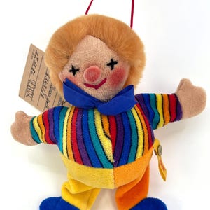 May include: A vintage clown doll with a plush, orange-yellow hair, rosy cheeks, and a blue bow tie. The doll wears a striped rainbow shirt, yellow and orange pants, and blue shoes. A small tag is attached to the doll.