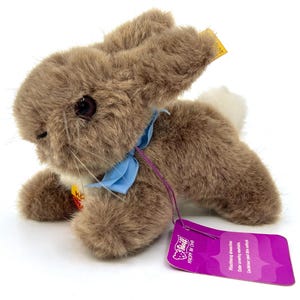 Puede incluir: Un conejo de peluche marrón con una cinta azul alrededor del cuello y una cola blanca. El conejo tiene ojos oscuros y bigotes. Una etiqueta púrpura está adjunta.