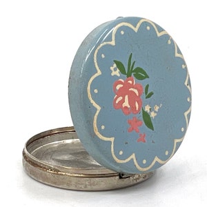 May include: A small, round, blue metal trinket box with a white scalloped edge and a floral design. The lid is open and the inside of the box is silver.