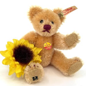Op de afbeelding: Een kleine, bruine teddybeer met een rode knoopneus en een zonnebloem in zijn poot. De beer heeft een Steiff-label aan zijn oor met de tekst "Original Knopf im Ohr".