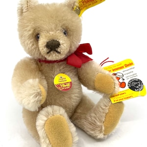 Peut inclure: Un ours en peluche beige Steiff avec un ruban rouge autour du cou. L'ours a un bouton sur la poitrine qui indique "Original Teddybär Steiff Knopf im Ohr". L'ours est assis sur un fond blanc.