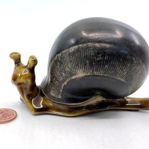 May include: A ceramic snail figurine with a brown and black shell and a brown body. The shell has a textured pattern.