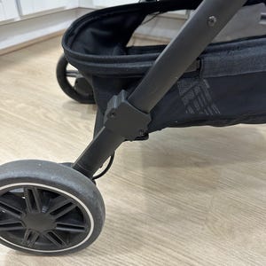 Puede incluir: Primer plano de un cochecito negro con un marco y ruedas negros. La rueda delantera es visible, con una llanta blanca y un neumático negro. También se ve la cesta del cochecito.