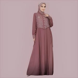 Op de afbeelding: Een lange mauve jurk met lange mouwen en een witte, gepatroneerde overlay en een knoopsluiting aan de voorkant. De jurk heeft een geplooide rok en een strik in de taille.