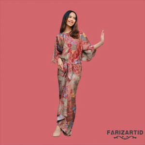 Puede incluir: Una mujer lleva un vestido largo y fluido con un estampado floral en tonos rojos, marrones y azules. El vestido tiene un lazo en la cintura y una silueta suelta y fluida. La mujer está de pie sobre un fondo rosa.