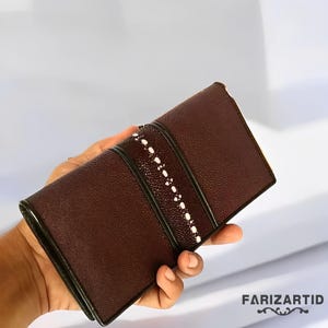 Könnte beinhalten: Eine braune Ledergeldbörse mit einem weißen und grünen Streifenmuster. Die Geldbörse hat eine strukturierte Oberfläche und ein weißes Nähdetail.