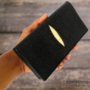 Könnte beinhalten: Schwarze Ledergeldbörse mit einem markanten weißen Stachelrochen-Design. Die Geldbörse ist rechteckig und scheint aus strukturiertem Leder zu bestehen. Die Geldbörse wird in einer Hand gehalten.