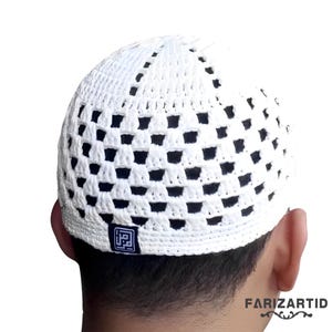 Puede incluir: Un gorro blanco de ganchillo con un logotipo cuadrado en un lado. El gorro tiene un diseño de patrón con pequeños agujeros en toda la superficie.