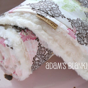 May include: A white faux fur baby blanket with a brown and pink floral print. The blanket is rolled up and has a tag that says "Adam's Blankie".