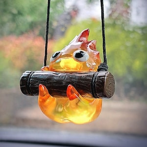 Op de afbeelding: Een geel en oranje glazen beeldje van een personage uit de film "Mijn buurman Totoro" hangend aan een zwart touwtje. Het personage houdt een bruine boomstam in zijn handen.