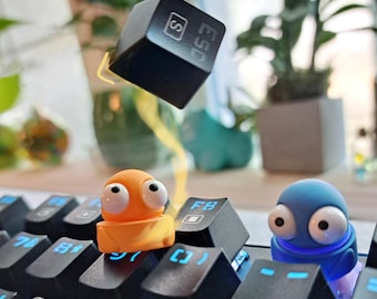 Può includere: Primo piano di una tastiera meccanica nera con tasti retroilluminati blu. L'immagine presenta tasti personalizzati, tra cui un tasto arancione e uno blu a forma di personaggio dei cartoni animati, e un tasto nero con la scritta "ESC" e un simbolo.