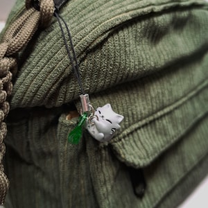 May include: Close-up of a green corduroy shoulder bag with a small gray cat charm hanging from the zipper. The bag has a flap pocket and a braided strap. The charm has a green tassel.