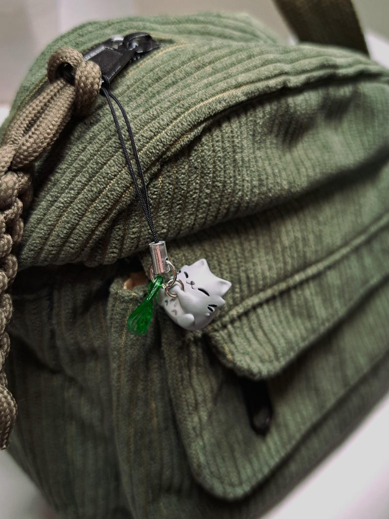 May include: Close-up of a green corduroy shoulder bag with a small gray cat charm hanging from the zipper. The bag has a flap pocket and a braided strap. The charm has a green tassel.