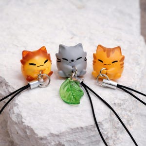 May include: Three cat-shaped charms in orange, yellow, and gray. Each charm has a black cord and a silver-colored clasp. One charm has a green leaf charm. The cat charms are small and have smiling faces.