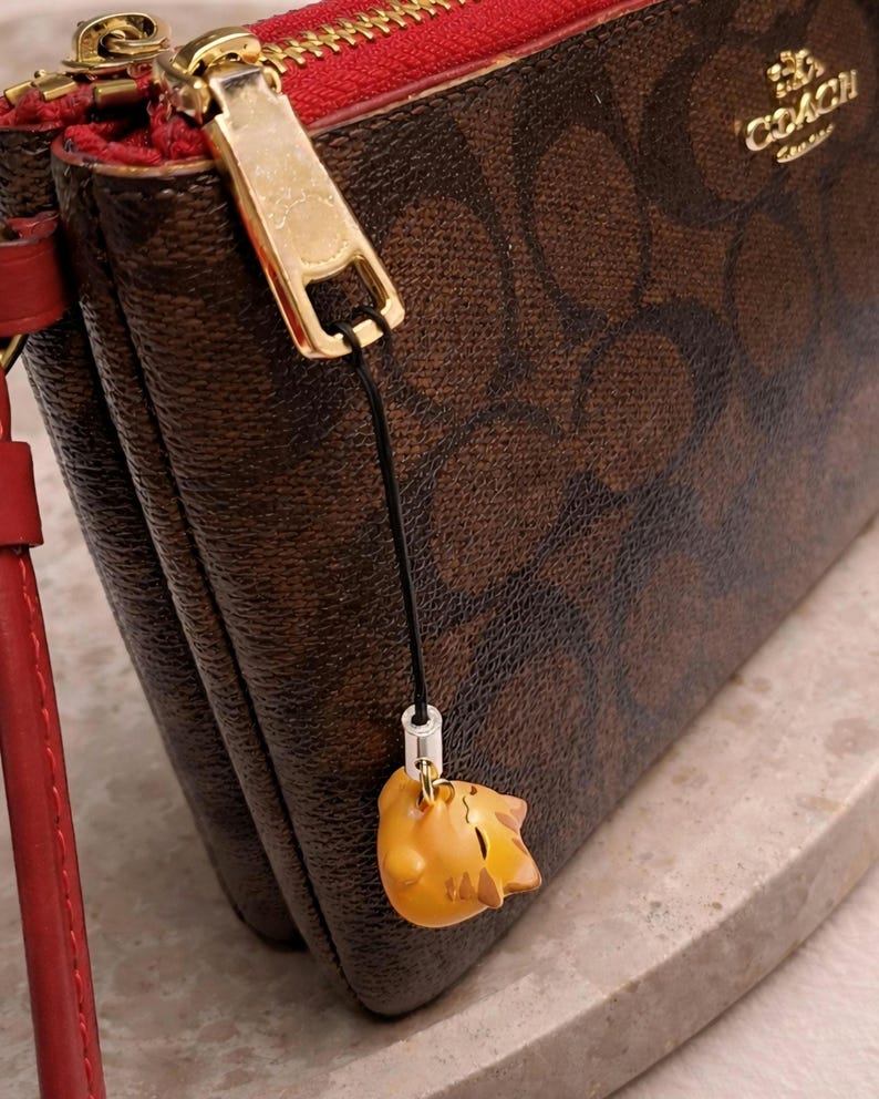 May include: Brown Coach wallet with a red interior and gold zipper. The wallet features a repeating pattern and a small, yellow cat charm hanging from the zipper. The Coach logo is visible in gold lettering.
