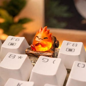 May include: A custom artisan keycap featuring a smiling, orange flame character resting on a piece of wood. The keycap is placed on a white keyboard, with the F6, F8, and other keys visible.