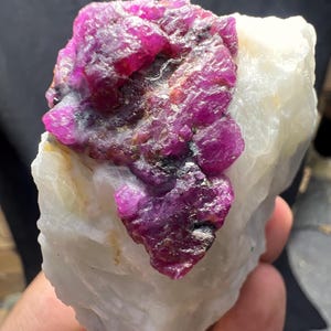 Puede incluir: Un grupo de cristales de rubí en bruto, con un vibrante tono magenta, se asienta sobre cuarzo blanco. La gema en bruto muestra una superficie texturizada, mostrando su estructura cristalina. Este espécimen mineral único es ideal para coleccionistas.