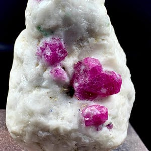 Puede incluir: Un espécimen mineral en bruto que presenta cristales de rubí de color rosa intenso incrustados en una matriz blanca opaca y texturizada. La piedra tiene una forma irregular y dentada con múltiples formaciones cristalinas distintas.