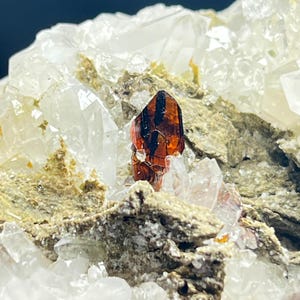 Puede incluir: Primer plano de un espécimen mineral con un cristal naranja oscuro con estrías negras, sobre una matriz de cristales de cuarzo transparente y una base texturizada de color marrón claro. Los cristales de cuarzo son translúcidos.