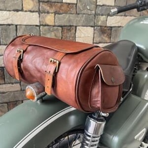 May include: A brown leather motorcycle bag is mounted on the rear of a green motorcycle. The bag is cylindrical with two buckle straps and a small side pocket. The motorcycle has a black tire and a silver suspension.