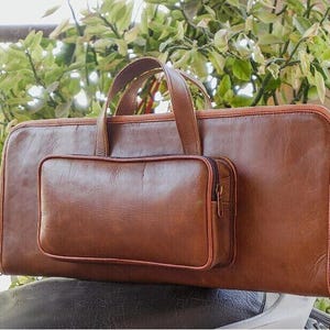 Peut inclure: Une mallette en cuir marron avec une petite pochette assortie attachée. La mallette a une poignée et une fermeture éclair. La pochette a une fermeture éclair. Le cuir a une texture lisse.