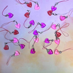 May include: A collection of heart-shaped charms in shades of pink and red, with silver-colored clasps and matching pink lanyards. The charms are scattered on a soft, gradient background, creating a playful and romantic aesthetic.