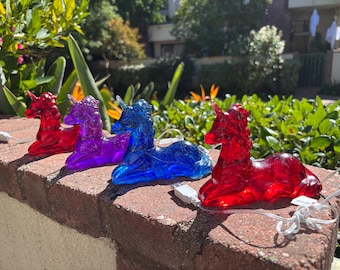 May include: Four colorful unicorn lights in red, purple, and blue sit on a brick wall. Each unicorn has a USB cord attached. The background features lush green foliage and a sunny outdoor setting.