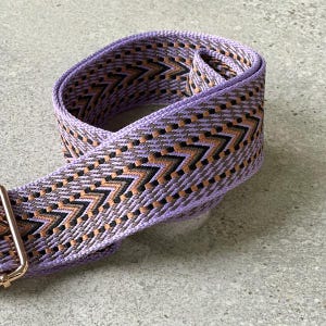 May include: A woven strap in shades of lavender, brown, and black, with a repeating chevron pattern. The strap has a gold-coloured metal clasp. The strap is designed for use with a bag or purse.