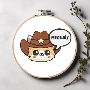 May include: Cross-stitch embroidery of a cartoon cat wearing a brown cowboy hat with a star. The cat has large eyes and a speech bubble that says "Meowdy". The embroidery is set in a wooden hoop.