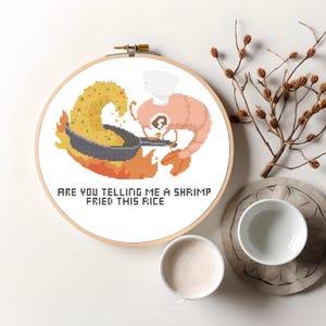 May include: A white embroidery hoop with a cross-stitch design of a shrimp wearing a chef's hat and cooking fried rice in a pan over a flame. The text "Are you telling me a shrimp fried this rice?" is below the design.
