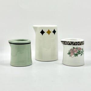 May include: Three ceramic cream pitchers. The pitcher on the left is light green with a black rim. The center pitcher is white with black and gold diamond shapes. The pitcher on the right is white with a floral design and black trim.