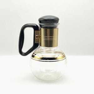 May include: A clear glass carafe with a black handle and lid, and a gold-colored band around the neck and base. The carafe has a rounded body and is designed for serving beverages.