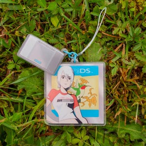 Peut inclure: Un porte-clés en acrylique transparent avec une illustration de personnage en chemise rouge et blanche et un colibri. Le porte-clés est attaché à une petite console de jeu portable argentée. Une lanière blanche est attachée au porte-clés.