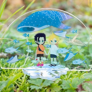Peut inclure: Une figurine en acrylique transparente de deux personnages de dessin animé debout devant un requin baleine bleu. Les personnages se tiennent la main et sont entourés de petits poissons bleus et d'une tortue de mer. La figurine est posée sur une base en acrylique transparente avec un motif de vague.