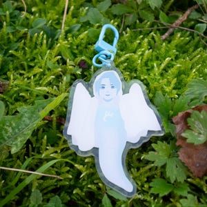 Peut inclure: Un porte-clés en acrylique transparent représentant une figure fantomatique bleu clair avec des ailes d'ange et un visage souriant. Il est attaché à un fermoir bleu clair. Le fond est de la mousse et du feuillage verts.