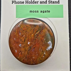 May include: A Rock Popup phone holder and stand featuring a moss agate stone. The holder is round, with a mix of brown, red, and white colors. The packaging includes the text "ROCK popup" and "Phone Holder and Stand."