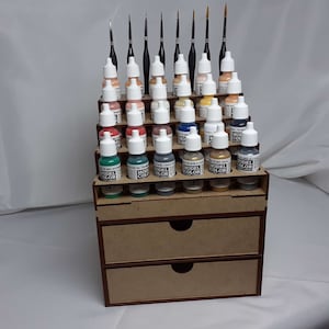 Puede incluir: Un estante de pintura de madera con tres cajones contiene filas de pequeños frascos de pintura acrílica. Los frascos están etiquetados con "Model Color" y están dispuestos en un patrón de arcoíris. El estante está hecho de madera marrón claro y tiene un borde marrón oscuro.
