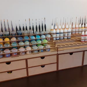 May include: A tiered wooden organizer featuring multiple circular cutouts for paint pots and vertical slots for brushes. The unit includes several drawers with circular finger pulls, providing storage for hobby supplies in a structured arrangement.