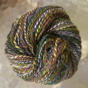 May include: A tightly wound ball of yarn in a variety of colors, including green, yellow, brown, and gray. The yarn appears to be a thick, textured blend, suitable for knitting or crocheting projects. The overall look is rustic and natural.