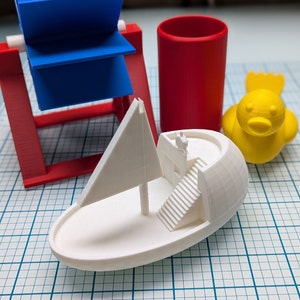 May include: A white 3D printed model of a sailboat with a sail and a staircase. The sailboat is sitting on a gridded surface. There is a red cylinder, a blue and red toy windmill, and a yellow toy duck in the background.