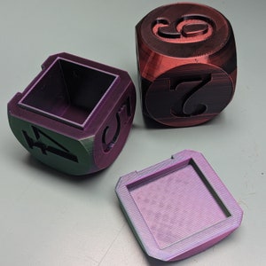 May include: Three 3D printed dice, one red, one purple, and one green. The dice are in the shape of a cube and have numbers on each side. The dice are sitting on a gray surface.