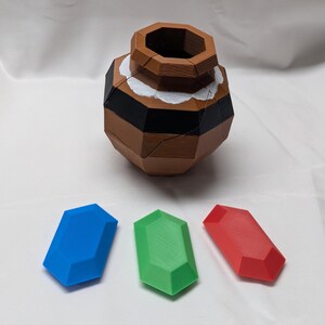 May include: A brown wooden puzzle in the shape of a pot with a white and black rim. Three gems are in front of the pot: a blue gem, a green gem, and a red gem.