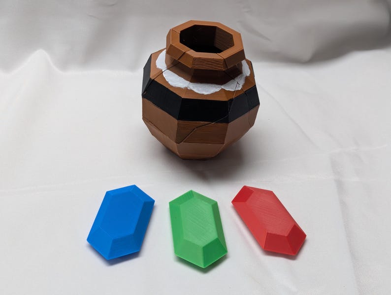 May include: A brown wooden puzzle in the shape of a pot with a white and black rim. Three gems are in front of the pot: a blue gem, a green gem, and a red gem.