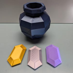 May include: Three 3D printed gems, one gold, one purple, and one silver, sitting on a gray surface in front of a dark blue, geometric vase.