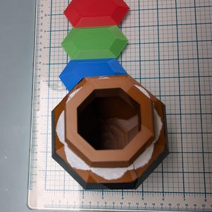 May include: A brown, octagonal, ceramic container with a white rim and a black base. The container has a large, open top. Three plastic gems, red, green, and blue, are arranged above the container.