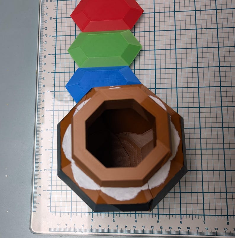 May include: A brown, octagonal, ceramic container with a white rim and a black base. The container has a large, open top. Three plastic gems, red, green, and blue, are arranged above the container.