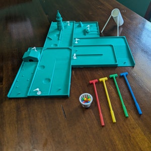 May include: A miniature golf course set with a green felt playing surface, a white plastic tower, a white plastic bucket, and five colorful plastic putters. The set includes a basket filled with colorful golf balls.