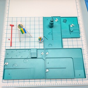May include: A miniature model of a playground with a blue and green color scheme. The playground features a slide, a swing set, a basketball hoop, and a small building. There are also some small white figures scattered around the playground.