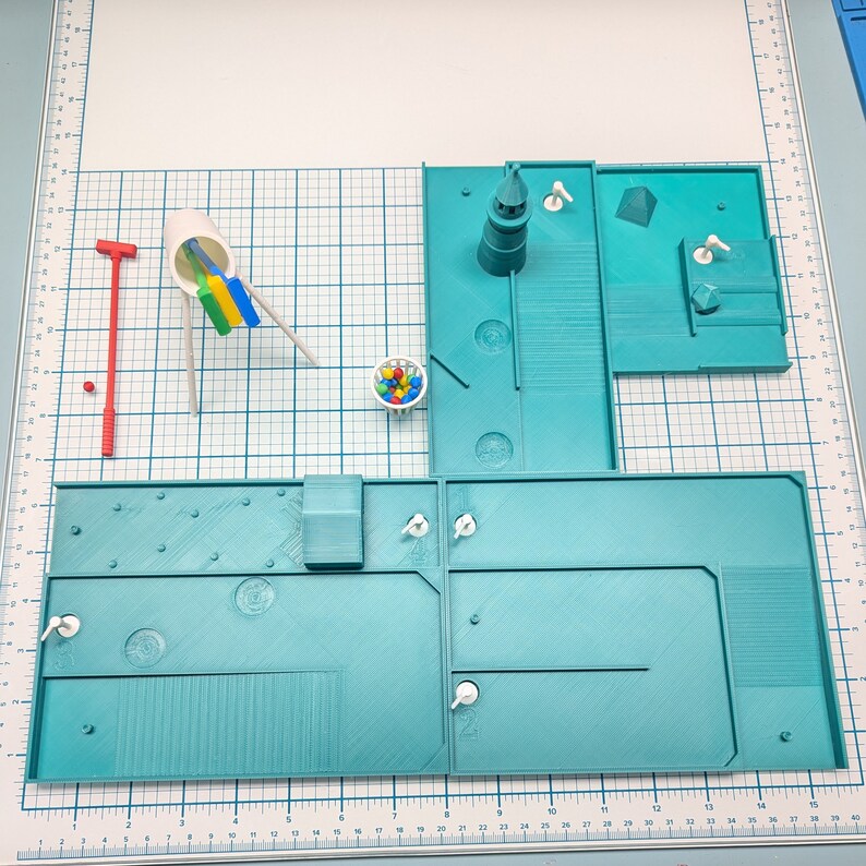 May include: A miniature model of a playground with a blue and green color scheme. The playground features a slide, a swing set, a basketball hoop, and a small building. There are also some small white figures scattered around the playground.