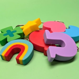 May include: A selection of brightly coloured keyrings in diverse shapes: a rainbow, a four-leaf clover, a star, a heart, a letter, and a curved arrow. Each keyring is attached to a silver metal ring.